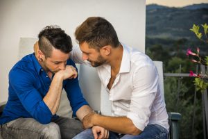 Dois homens estão sentados num pátio, um vestido com uma camisa azul e outro com uma camisa branca. O homem de azul, perdido nas suas inseguranças, descansa a cabeça na mão enquanto o outro segura gentilmente no braço, transmitindo apoio para o ajudar a vencer os seus medos.