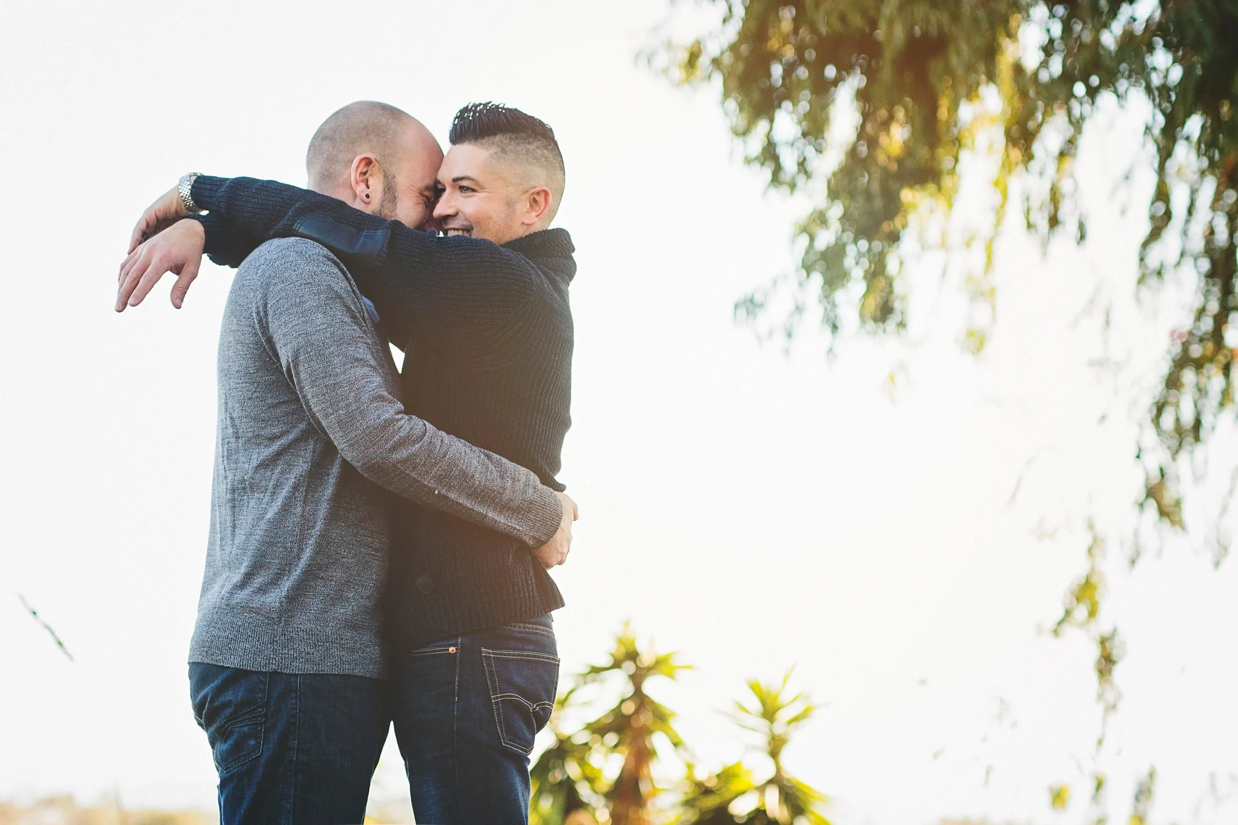 Duas pessoas abraçam-se ao ar livre, ambas sorriem e usam camisas de manga compridas. Árvores e plantas são visíveis ao fundo, enquanto ponderam como contar aos amigos sobre sua jornada juntos.