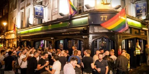 Uma multidão de pessoas reúne-se à noite em frente ao pub "The Duke of Wellington", um dos melhores bares de Londres. Bandeiras arco-íris pendentes do exterior do edifício, celebrando a sua vibrante comunidade gay.
