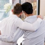 Duas pessoas de costas para a câmara sentam-se próximas, com os braços à volta uma da outra, numa sala iluminada com grandes janelas, revelando um momento terno de amor e ligação em meio aos problemas da vida.