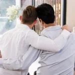 Duas pessoas de costas para a câmara sentam-se próximas, com os braços à volta uma da outra, numa sala iluminada com grandes janelas, revelando um momento terno de amor e ligação em meio aos problemas da vida.