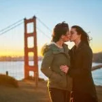 Duas pessoas com casacos compartilharam um beijo ao pôr do sol em frente à Ponte Golden Gate, capturando um momento perfeito que certamente receberá muitos gostos no Facebook.