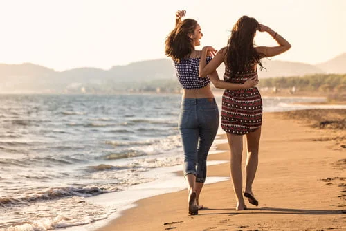 Duas mulheres, uma de calças de ganga e a outra com um vestido, caminham de braço dado ao longo de uma praia arenosa junto ao oceano, com colinas visíveis ao fundo. A sua alegria e ligação ilustram as vantagens simples mas profundas do amor, celebrando o seu romance homossexual contra o deslumbrante cenário natural.