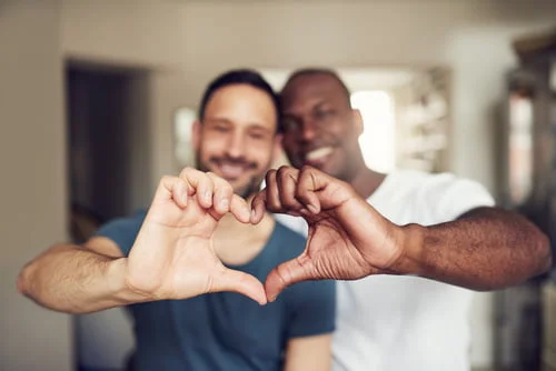 Duas pessoas estão de pé, próximas uma da outra, sorrindo e formando um coração com as mãos, como se estiverem prontas para contar aos amigos sobre sua orientação sexual.