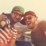 Dois homens sorridentes, vestidos com roupas casuais, óculos de sol e bonés, tiraram uma selfie ao ar livre com um smartphone, destacando as vantagens de sair com um gay em sua aventura divertida.