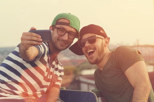 Dois homens sorridentes, vestidos com roupas casuais, óculos de sol e bonés, tiraram uma selfie ao ar livre com um smartphone, destacando as vantagens de sair com um gay em sua aventura divertida.