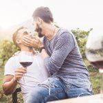 Dois homens estão sentados juntos numa vinha, um segurar um copo de vinho tinto e o outro a acariciar-lhe o rosto com ternura. Ambos sorriem, desfrutando do seu tempo num dos belos destinos gays europeus, com copos de vinho e verdes vinhas exuberantes ao fundo.