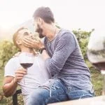 Dois homens estão sentados juntos numa vinha, um segurar um copo de vinho tinto e o outro a acariciar-lhe o rosto com ternura. Ambos sorriem, desfrutando do seu tempo num dos belos destinos gays europeus, com copos de vinho e verdes vinhas exuberantes ao fundo.