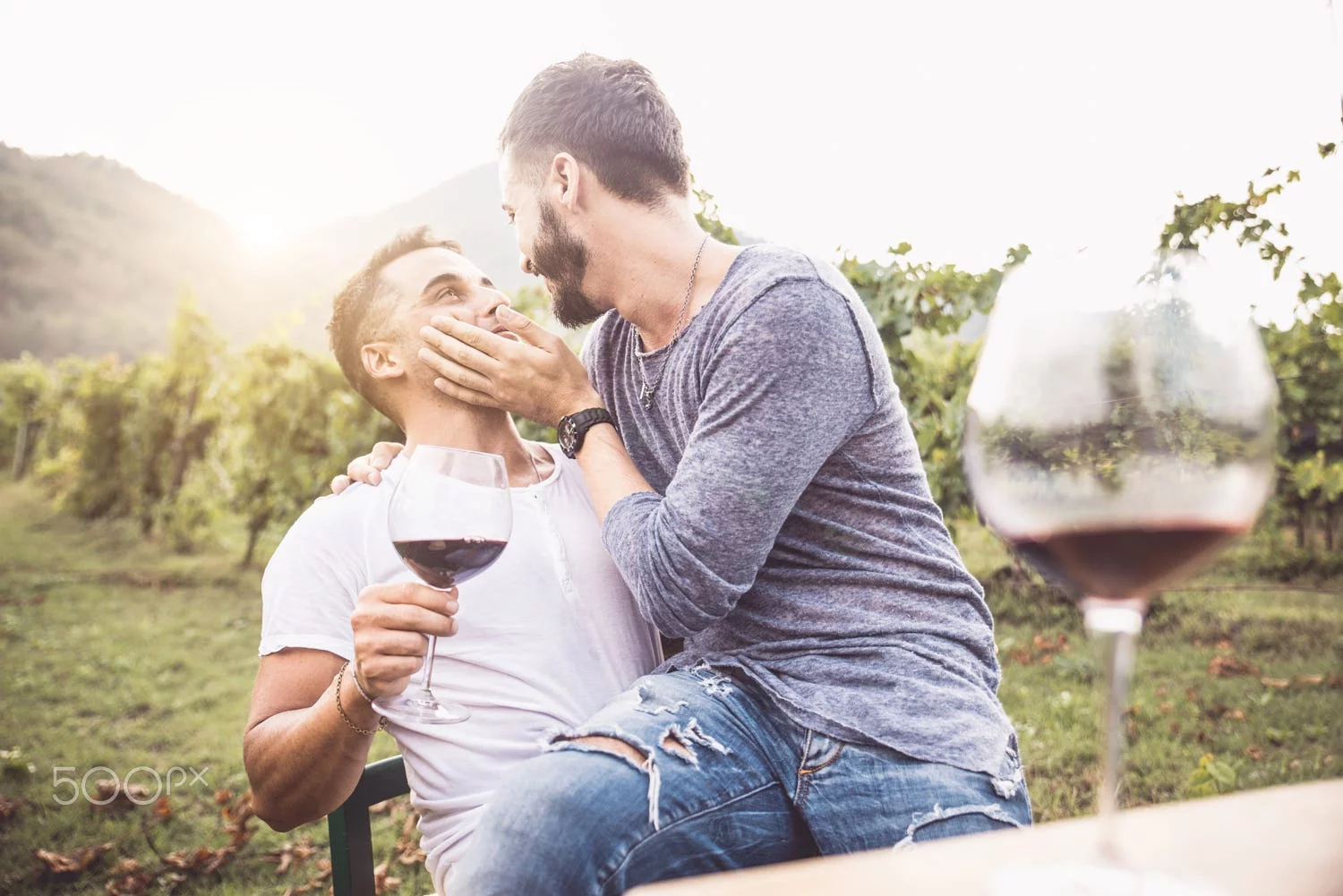 Dois homens estão sentados juntos numa vinha, um segurar um copo de vinho tinto e o outro a acariciar-lhe o rosto com ternura. Ambos sorriem, desfrutando do seu tempo num dos belos destinos gays europeus, com copos de vinho e verdes vinhas exuberantes ao fundo.