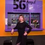 A mulher trans num vestido preto está em frente ao balcão do bar no clube "Apolo 56", com garrafas de licor dispostas nas prateleiras atrás dela e um ramo de flores no balcão.