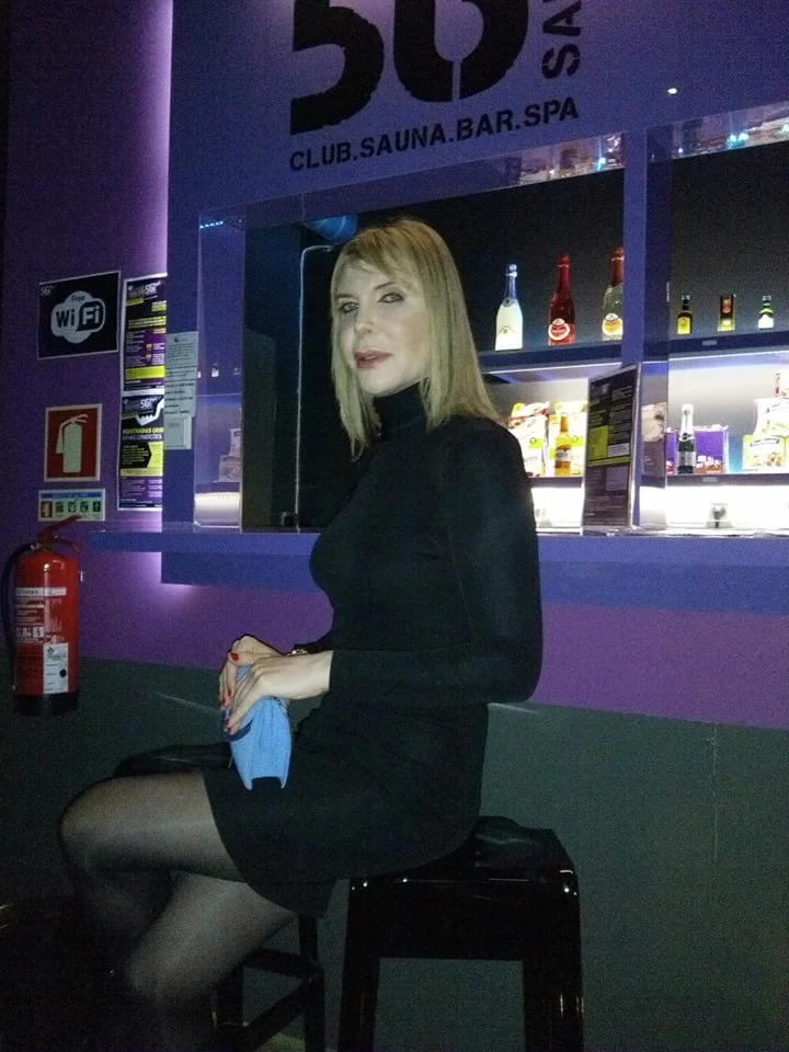 A mulher trans usa um vestido preto e meias está sentada num banquinho em frente ao balcão do bar, com garrafas e um espelho atrás dela. O ambiente parece ser um clube com iluminação fraca e paredes roxas, heteroamigável e acolhedor para todos.