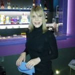 Uma mulher trans de cabelo loiro, usa um vestido preto e segura uma clutch azul, sorri enquanto posa em frente a um bar com várias garrafas de licor.