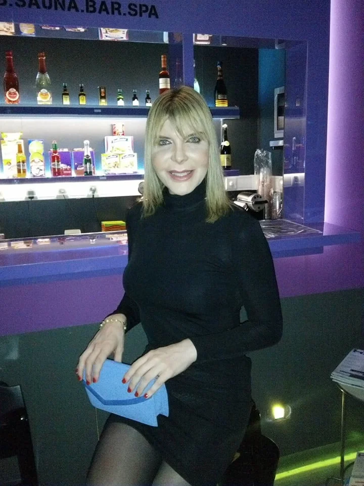 Uma mulher trans de cabelo loiro, usa um vestido preto e segura uma clutch azul, sorri enquanto posa em frente a um bar com várias garrafas de licor.