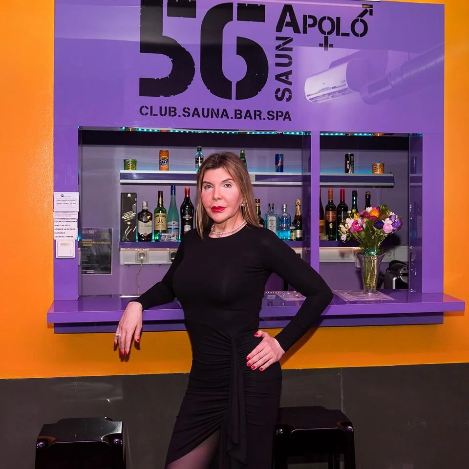 Uma mulher com um vestido preto está em frente a uma bancada de bar no interior de um clube chamado "56 Sauna Apolo". O bar apresenta várias garrafas de álcool e um vaso com flores. Paredes roxas e amarelas enquadraram a cena, criando o cenário para Kiki Pais de Sousa, cuja história de vida como trans portuguesa continua a inspirar.