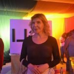 A mulher trans portuguesa, Kiki Pais de Sousa, encontra-se num espaço interior sob uma iluminação colorida, com o cabelo comprido solto e vestidos uma camisola preta. Outras pessoas e um grande painel branco com letras pretas são visíveis ao fundo, acrescentando camadas à sua história de vida.