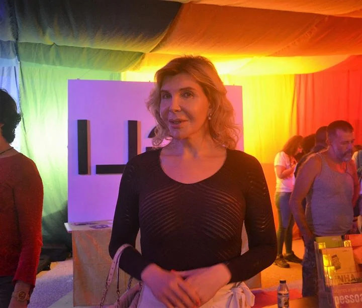 A mulher trans portuguesa, Kiki Pais de Sousa, encontra-se num espaço interior sob uma iluminação colorida, com o cabelo comprido solto e vestidos uma camisola preta. Outras pessoas e um grande painel branco com letras pretas são visíveis ao fundo, acrescentando camadas à sua história de vida.