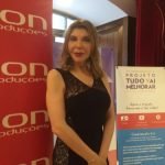 Uma pessoa vestida de preto está em um ambiente interno, ao lado de cartazes, um dos quais diz: "Projeto Tudo Vai Melhorar". O cenário parece ser um evento formal ou promocional, promovendo apoio a todas as meninas e meninos.