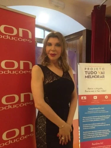 Uma pessoa vestida de preto está em um ambiente interno, ao lado de cartazes, um dos quais diz: "Projeto Tudo Vai Melhorar". O cenário parece ser um evento formal ou promocional, promovendo apoio a todas as meninas e meninos.