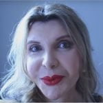 Uma mulher transexual com cabelo loiro comprido e batom vermelho olha para a câmara. Tem uma expressão neutra e está numa sala com uma cortina escura e paredes de cor cinza-claro.