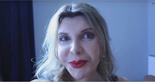 Uma mulher transexual com cabelo loiro comprido e batom vermelho olha para a câmara. Tem uma expressão neutra e está numa sala com uma cortina escura e paredes de cor cinza-claro.