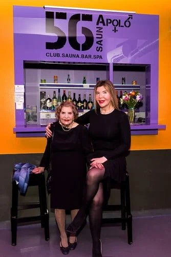 Duas mulheres vestidas de preto sentaram-se em bancos altos em frente a um balcão de bar com várias garrafas. Um letreiro acima diz "56 Apolo Club. Sauna. Bar. Spa." A parede ao fundo é amarela e roxa. A rapariga serve bebidas discretamente, contribuindo para uma atmosfera vibrante.