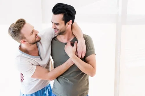 Dois homens estão juntos, sorrindo e abraçados, com um braço sobre os ombros um do outro. O fundo é suavemente iluminado com luz natural, criando uma atmosfera acolhedora que desperta curiosidade sobre a sua ligação.