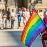 Pessoa segurando uma bandeira arco-íris enquanto usa uma mochila vermelha, com uma multidão desfocada ao fundo num dia solarengo, a celebrar o orgulho num dos muitos países a visitar para uma experiência inclusiva e gay-friendly.