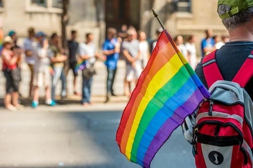 Pessoa segurando uma bandeira arco-íris enquanto usa uma mochila vermelha, com uma multidão desfocada ao fundo num dia solarengo, a celebrar o orgulho num dos muitos países a visitar para uma experiência inclusiva e gay-friendly.