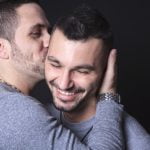 Dois homens abraçam-se, um a beijar a bochecha do outro. Pedro e seu companheiro, ambos vestidos com camisas cinzentas, sorriem e demonstram afeto contra um fundo preto. Este momento destaca a normalidade de sua história de amor como casal homossexual.