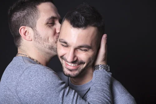 Dois homens abraçam-se, um a beijar a bochecha do outro. Pedro e seu companheiro, ambos vestidos com camisas cinzentas, sorriem e demonstram afeto contra um fundo preto. Este momento destaca a normalidade de sua história de amor como casal homossexual.