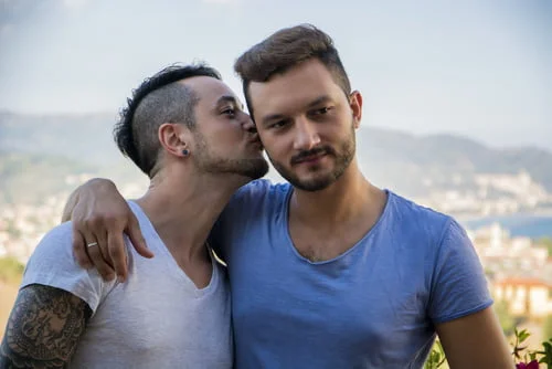 Dois homens em pé ao ar livre, com um a beijar o outro na cara. Ambos vestem camisolas casuais e sorriem preferencialmente. Os melhores amigos gays a compartilhar um momento especial sem precisarem de explicação.