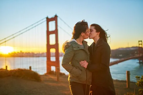 Duas pessoas estão a beijar-se em frente à Ponte Golden Gate ao pôr-do-sol, com uma a usar um casaco verde e a outra um casaco preto. Este momento íntimo destaca a beleza e inclusividade do amor LGBTQ+ num cenário tão pitoresco.