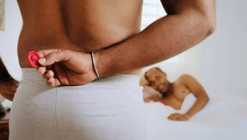 Um homem com calções cinzentas, segurando um preservativo vermelho atrás das costas, enquanto outro homem está deitado na cama ao fundo. Curiosidades sobre sexo gay são sutilmente sugeridas nesta cena intrigante.