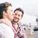 Dois homens, claramente amigos gays, sentam-se juntos num banco, um com o braço à volta do outro, sorrindo e olhando um para o outro. Ao fundo, vê-se um cenário desfocado ao ar livre com pessoas e estruturas.