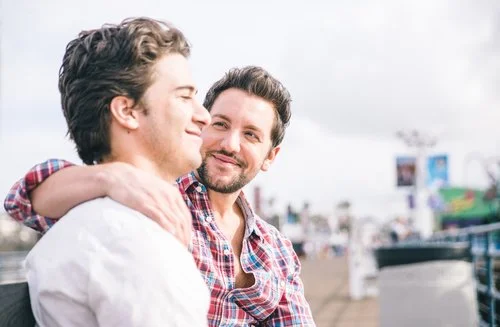 Dois homens, claramente amigos gays, sentam-se juntos num banco, um com o braço à volta do outro, sorrindo e olhando um para o outro. Ao fundo, vê-se um cenário desfocado ao ar livre com pessoas e estruturas.