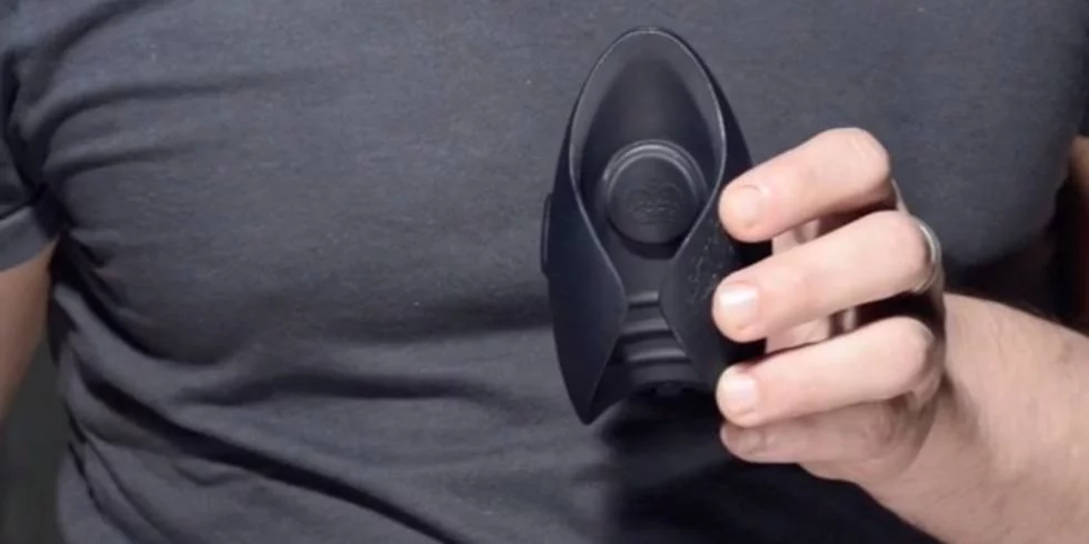 Uma pessoa com uma camisa escura segura um pequeno dispositivo oval preto com uma mão, possivelmente um cara discreto.