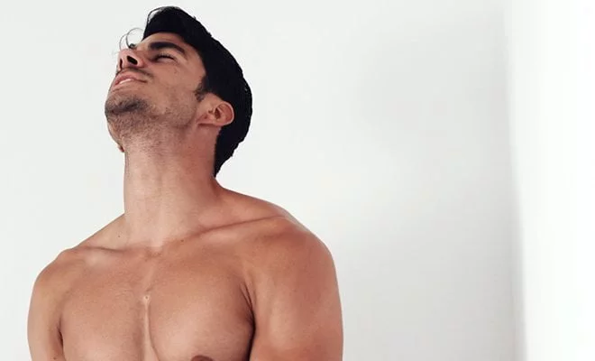 Pessoa sem camisa, de cabelo escuro e barba por fazer, inclina a cabeça para trás, olhos fechados, contra um fundo branco, segurando um Guybrator na mão.