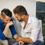 Dois homens, celebrando a sua homossexualidade, estão sentados juntos num deque ao ar livre. Um está a beijar a mão do outro enquanto se dá as mãos e se olha. São rodeados pela natureza e por algumas flores.