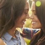 Duas mulheres de cabelo comprido, a sorrir frente a frente, tocando com as testas e abraçando-se num cenário ao ar livre iluminado pelo sol, criando um verdadeiro espetáculo de amor.