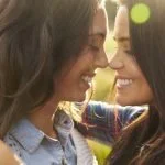 Duas mulheres de cabelo comprido, a sorrir frente a frente, tocando com as testas e abraçando-se num cenário ao ar livre iluminado pelo sol, criando um verdadeiro espetáculo de amor.