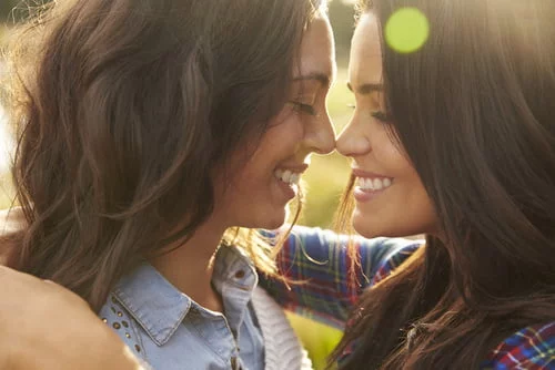Duas mulheres de cabelo comprido, a sorrir frente a frente, tocando com as testas e abraçando-se num cenário ao ar livre iluminado pelo sol, criando um verdadeiro espetáculo de amor.