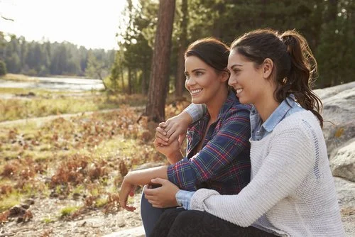 Duas mulheres, que poderiam ser namoradas, estão sentadas próximas numa grande rocha ao ar livre, sorrindo e olhando para o horizonte. As árvores frondosas e a natureza em seu redor criam um espetáculo digno de admiração.