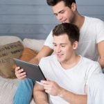 Dois jovens em roupas informais sentados no sofá, um braço um tablet e o outro a olhar por cima do ombro, possivelmente a navegar pelo Dicionário LGBT.