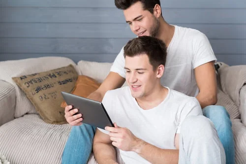 Dois jovens em roupas informais sentados no sofá, um braço um tablet e o outro a olhar por cima do ombro, possivelmente a navegar pelo Dicionário LGBT.