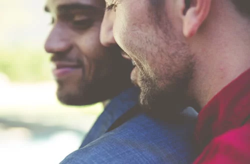 Close de dois homens heterossexuais de pé lado a lado, ambos sorrindo. Um deles veste um casaco azul, ou outro um casaco vermelho. As suas faces estão progressivamente viradas para o lado, com um fundo desfocado e brilhante que confere um toque de fantasia.