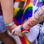 Duas pessoas de mãos dadas, a usar acessórios arco-íris e uma bandeira do orgulho. Uma das pessoas tem "ORGULHO!" pintado no braço, destacando a importância da visibilidade, visto que a homossexualidade continua ilegal em 72 países.