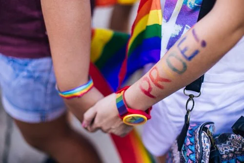 Duas pessoas de mãos dadas, a usar acessórios arco-íris e uma bandeira do orgulho. Uma das pessoas tem "ORGULHO!" pintado no braço, destacando a importância da visibilidade, visto que a homossexualidade continua ilegal em 72 países.