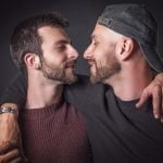 Dois homens com barba, um a usar uma camisola de malha cor de vinho e o outro a usar um boné preto e uma camisa escura, estão frente a frente, abraçados, contra um fundo escuro, exemplificando como se pode combater a homofobia através da solidariedade e fazer afeto.