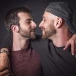 Dois homens com barba, um a usar uma camisola de malha cor de vinho e o outro a usar um boné preto e uma camisa escura, estão frente a frente, abraçados, contra um fundo escuro, exemplificando como se pode combater a homofobia através da solidariedade e fazer afeto.
