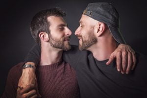 Dois homens com barba, um a usar uma camisola de malha cor de vinho e o outro a usar um boné preto e uma camisa escura, estão frente a frente, abraçados, contra um fundo escuro, exemplificando como se pode combater a homofobia através da solidariedade e fazer afeto.